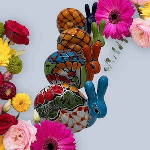 May include: A stack of four ceramic snail figurines with long ears, each featuring distinct hand-painted patterns in vibrant colors including orange, blue, red, and green, surrounded by a colorful arrangement of fresh pink, red, and yellow flowers.