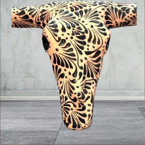 May include: A decorative cow skull featuring a cream base and detailed black floral patterns. The skull has a smooth, glossy finish, designed for wall mounting. The symmetrical design covers the entire surface, offering a striking visual impact.
