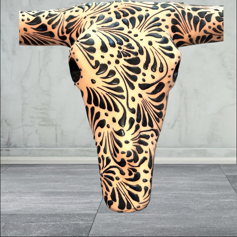 May include: A decorative cow skull featuring a cream base and detailed black floral patterns. The skull has a smooth, glossy finish, designed for wall mounting. The symmetrical design covers the entire surface, offering a striking visual impact.
