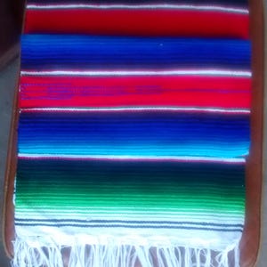 K&ouml;nnte beinhalten: Eine bunte mexikanische Decke mit wei&szlig;en Fransen. Die Decke hat Streifen in Rot, Gr&uuml;n, Blau, Gelb und Wei&szlig;.