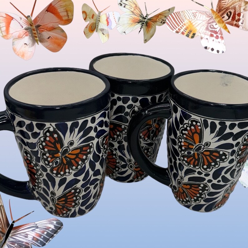 May include: Three ceramic mugs with blue and white butterfly designs. The mugs have a dark blue rim and a white background with blue and white floral patterns. The butterflies are orange and black.