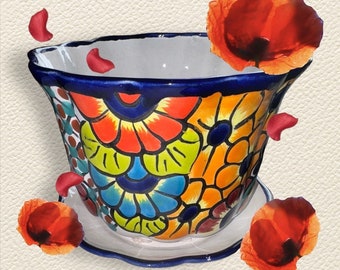 May include: A colorful ceramic planter with a blue rim and a floral design. The planter has a scalloped edge and is sitting on a white surface. There are red flower petals scattered around the planter.