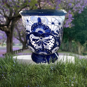 May include: A large, blue and white ceramic planter with a detailed floral design. The planter has a wide, slightly flared rim and a pedestal base. The intricate pattern features stylized flowers and leaves. The planter is displayed on a white pedestal in a garden setting.