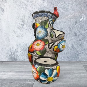 Puede incluir: Una maceta alta de cer&aacute;mica pintada a mano con un acabado gris texturizado. La maceta presenta dise&ntilde;os florales coloridos en rojo, azul y amarillo, con un peque&ntilde;o p&aacute;jaro rojo posado en la parte superior. Tiene m&uacute;ltiples aberturas para plantas.