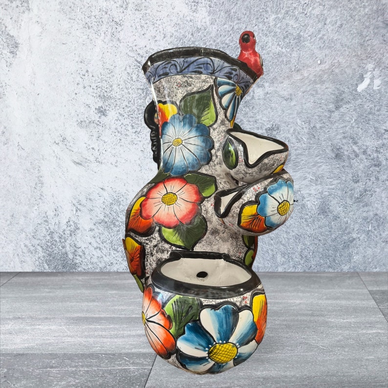 Puede incluir: Una maceta alta de cer&aacute;mica pintada a mano con un acabado gris texturizado. La maceta presenta dise&ntilde;os florales coloridos en rojo, azul y amarillo, con un peque&ntilde;o p&aacute;jaro rojo posado en la parte superior. Tiene m&uacute;ltiples aberturas para plantas.