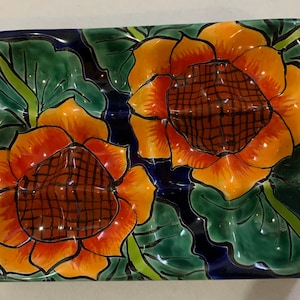 May include: A ceramic serving dish with a blue background and two orange and red sunflowers. The dish is decorated with green leaves and has a glossy finish.