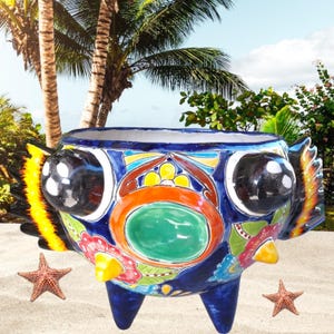 May include: A colourful ceramic planter shaped like a fish with large eyes, a green circle in the centre, and yellow, orange, red, blue and white details. The planter has three legs and is sitting on a sandy beach.