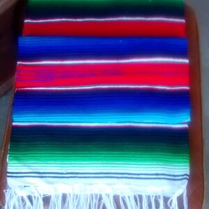 K&ouml;nnte beinhalten: Eine bunte mexikanische Decke mit wei&szlig;en Fransen. Die Decke hat Streifen in Rot, Blau, Gr&uuml;n, Gelb und Wei&szlig;.