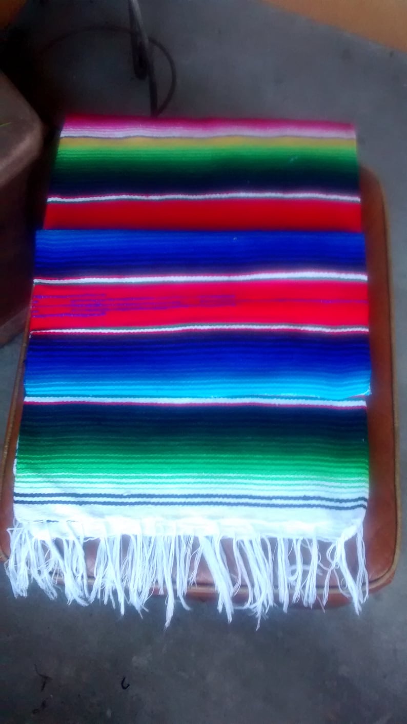 K&ouml;nnte beinhalten: Eine bunte mexikanische Decke mit wei&szlig;en Fransen. Die Decke hat Streifen in Rot, Blau, Gr&uuml;n, Gelb und Wei&szlig;.