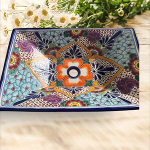 May include: Rectangular ceramic serving dish with a vibrant floral design. The dish features a blue border and a central flower in orange, white, and blue. The background is a mix of blue, teal, and patterned details, creating a colorful and decorative piece.
