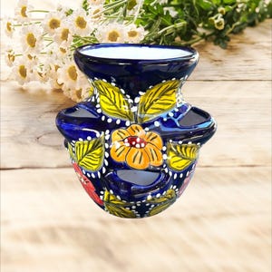Puede incluir: Un jarrón de cerámica azul cobalto pintado a mano con un diseño floral. El jarrón presenta una flor amarilla y naranja, hojas verdes y amarillas y detalles de puntos blancos. El jarrón tiene una forma única con dos asas laterales.