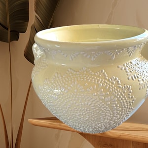 May include: A pale yellow ceramic planter with a white embossed design. The planter has two handles on the sides and a wide opening at the top.
