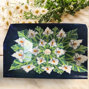 May include: A rectangular, navy blue ceramic serving dish with a floral design. The design features white calla lilies with orange centers and green leaves. The dish is likely used for serving food or as a decorative piece.