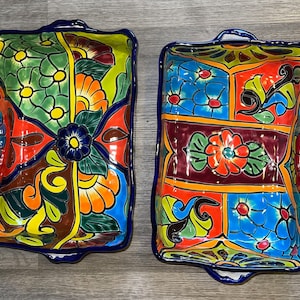 Pode incluir: Duas travessas retangulares de cerâmica com vibrantes desenhos florais. As travessas apresentam uma borda e alças azuis, com uma paleta colorida de laranja, verde, amarelo e azul. Os intrincados padrões lembram a cerâmica tradicional mexicana.