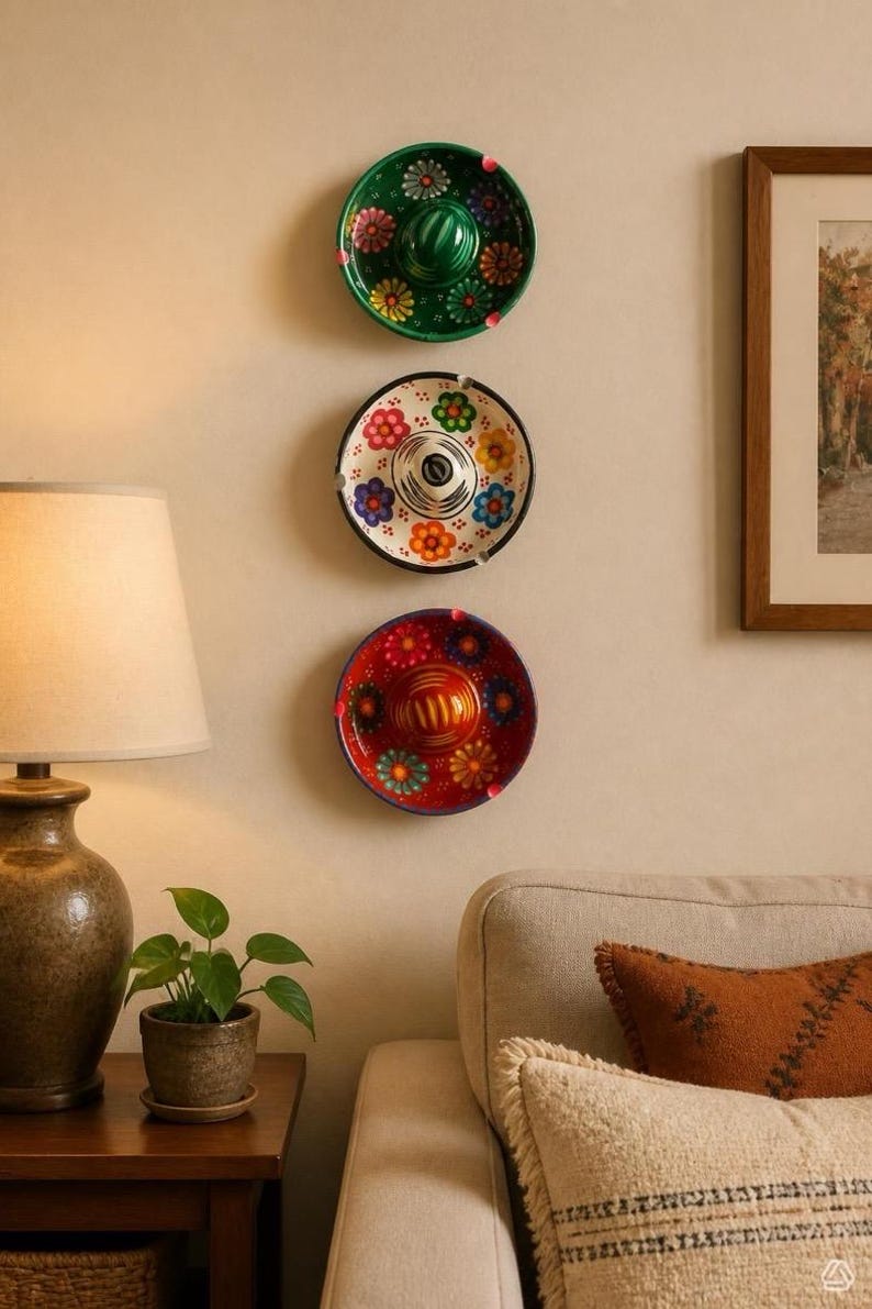 May include: Three circular ceramic ashtrays shaped like miniature sombreros are mounted vertically on a wall. The top one is green, the middle is white with a black rim, and the bottom is red. Each features colorful painted floral patterns.