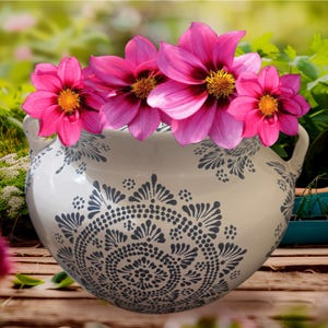 Op de afbeelding: Een witte keramische kom met twee handvatten aan de zijkant, versierd met een donkergrijs mandala-patroon van stippen en bloemblaadjes. De kom is gevuld met vier felroze dahlia's, elk met een helder geel hart en donkerpaarse kleurnuances.