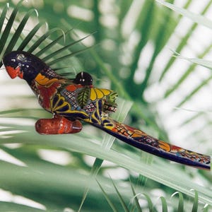 May include: A colorful ceramic parrot figurine with a detailed design. The parrot features a black head, red beak, and a body adorned with yellow, orange, and blue patterns. The figurine is perched on a brown base, set against a backdrop of green foliage.