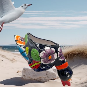 May include: A hand-painted ceramic planter shaped like a bird with colorful floral designs. The planter is black with purple, orange, yellow, green, and white flowers. The planter is sitting on a white rock.