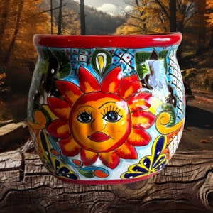 May include: A colourful ceramic planter with a red rim and a painted design of a yellow sun with a smiling face and red lips. The sun is surrounded by blue, green and red flowers and leaves.
