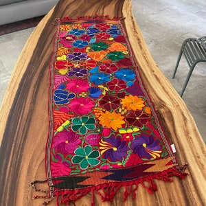 Puede incluir: Un camino de mesa bordado colorido con un patrón floral en tonos de rojo, rosa, naranja, amarillo, verde, azul y morado. El camino de mesa tiene un borde de flecos y está hecho de una tela suave y tejida.