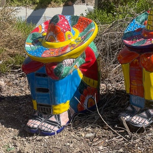 May include: Three brightly coloured ceramic figures of people wearing sombreros. The figures are painted with vibrant colours, including red, blue, yellow, and green. They are in a seated position, with their heads bowed and their arms resting on their knees. The figures are wearing sandals.