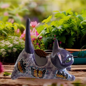 May include: A ceramic cat-shaped planter in shades of purple and gray, adorned with butterfly designs. The cat has green eyes and a tall, upright tail. The planter is designed to hold small plants or flowers, adding a whimsical touch to any garden or indoor space.
