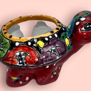 May include: A hand-painted ceramic turtle planter with a colourful floral design. The planter is red with green, yellow, purple, and white accents. It has a round opening on the top for planting.