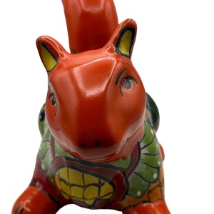 May include: A hand-painted ceramic squirrel figurine with bright orange, green, yellow, and red colors. The squirrel is sitting on its haunches with its tail curled up behind it.