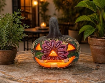 Può includere: Una lanterna di zucca intagliata con un disegno floreale dipinto nei toni del viola, verde e bianco. La zucca presenta occhi triangolari e un'ampia apertura dentellata per la bocca, illuminata dall'interno da una luce calda.