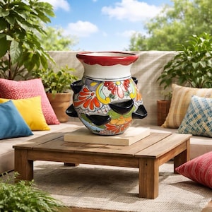 May include: A ceramic strawberry planter featuring a hand-painted floral pattern in vibrant shades of red, orange, yellow, and green. The pot has a wide white rim with a red edge and multiple dark blue planting pockets around its rounded body.