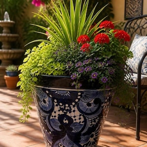 May include: A large, tapered ceramic planter featuring a glossy dark blue and white hand-painted floral pattern. The planter is filled with a vibrant arrangement of tall green spiky foliage, bright red geraniums, and small purple flowers.
