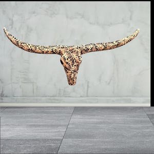 May include: A decorative steer skull with long horns. The skull is tan with a black floral pattern. The horns curve outwards. The skull is mounted on a grey wall.
