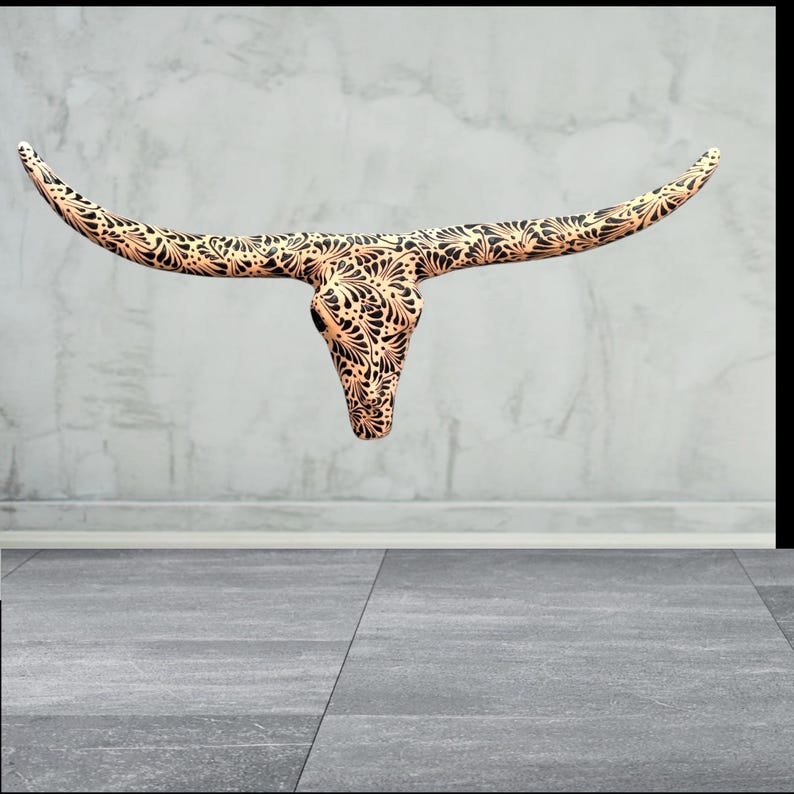 May include: A decorative steer skull with long horns. The skull is tan with a black floral pattern. The horns curve outwards. The skull is mounted on a grey wall.