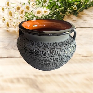 Puede incluir: Una olla de cerámica gris con interior marrón oscuro y borde negro. La olla presenta un patrón floral repetido tallado en el exterior y dos asas pequeñas. Flores blancas y vegetación en el fondo.