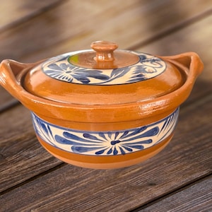 Puede incluir: Una cazuela de cerámica de color terracota con tapa. El plato presenta patrones florales azules y blancos alrededor de la base y en la tapa. Tiene dos asas y un pequeño pomo en la parte superior de la tapa.