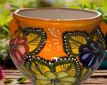May include: A vibrant orange ceramic planter with a glossy finish. The planter is adorned with hand-painted floral designs in red, blue, and yellow, set against green leaves. It has two small handles and a neutral interior, perfect for displaying plants.