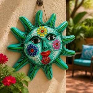 May include: A turquoise ceramic sun wall plaque featuring a stylised face with red lips, black eyes, and painted floral details in yellow, pink, and blue. The sun has multiple radiating points and is suspended by a thin brown twine cord.
