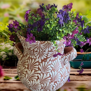 May include: A terracotta planter with two side handles, featuring a repeating white painted fan-like pattern across its exterior. The planter is filled with a dense arrangement of small purple flowers and green foliage, creating a vibrant contrast.