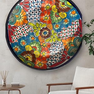 May include: A large, round, decorative ceramic plate with a vibrant floral design. The plate features a dark blue rim and a colorful pattern of flowers in shades of red, orange, yellow, green, and blue. The plate is mounted on a light-colored wall.