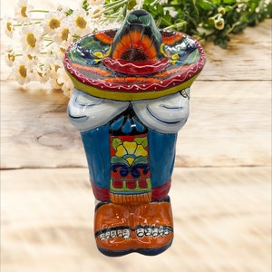 May include: A colourful ceramic figurine of a person wearing a sombrero and traditional Mexican clothing. The figurine is painted with bright colours, including blue, yellow, red and green.
