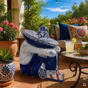 May include: A ceramic sculpture of a person sitting with knees tucked, wearing a wide-brimmed sombrero and pants, decorated in a blue and white hand-painted pattern. The figure is positioned on a tiled patio floor near potted plants and furniture.