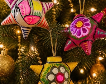 May include: Three star-shaped ornaments hanging from gold strings. The top left ornament is cream with pink, yellow, and green painted details. The top right is magenta with a purple flower design. The bottom is yellow with a white and pink flower.