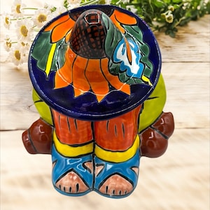 May include: A colorful ceramic figurine of a person wearing a sombrero with a sunflower design. The figurine is sitting with its legs crossed and has brown shoes.