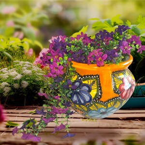 May include: An orange ceramic flower pot with a floral design, filled with purple and pink flowers. The pot has a wide opening and is decorated with blue, yellow, and red flowers. The flowers cascade over the side of the pot, creating a vibrant display.