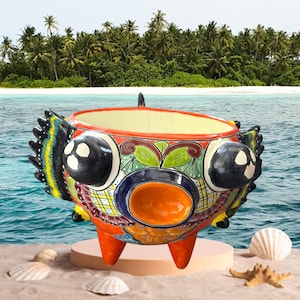 May include: A vibrant, hand-painted ceramic planter with a fish-like design. The planter features a colorful array of patterns and hues, including orange, blue, yellow, and green. It has three legs and a wide opening, perfect for displaying plants. The background shows a beach scene.
