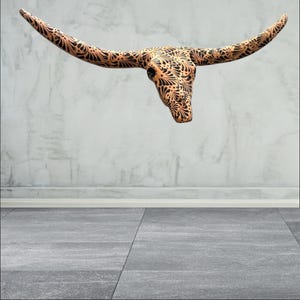 May include: A decorative steer skull with long, curved horns. The skull is a light brown colour with intricate black floral patterns covering the surface. The skull is mounted on a grey wall.