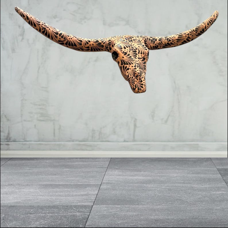 May include: A decorative steer skull with long, curved horns. The skull is a light brown colour with intricate black floral patterns covering the surface. The skull is mounted on a grey wall.