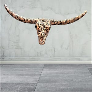 May include: A decorative steer skull with long horns, featuring a copper-coloured base and intricate black floral patterns. The skull is mounted on a wall, showcasing a rustic yet elegant design. The horns extend outwards, adding to its striking appearance.