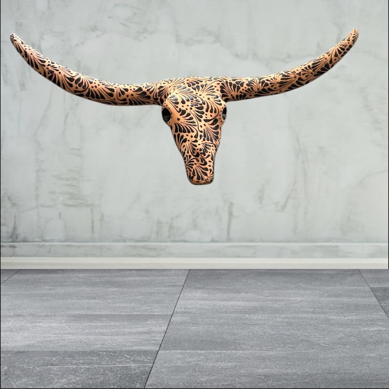 May include: A copper-toned longhorn skull featuring detailed black floral patterns. The skull is mounted on a wall, highlighting its decorative design. The horns curve outwards, creating a striking visual impact. The overall aesthetic is rustic and detailed.
