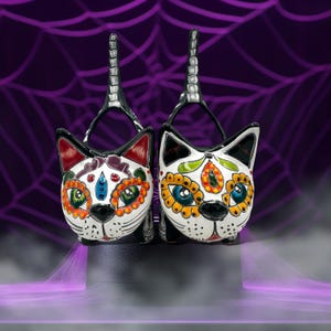 May include: Two decorative cat-shaped purses with Day of the Dead-inspired designs. The white ceramic cat faces feature colorful floral and geometric patterns, black ears, and a black nose. The purses have silver handles and are displayed against a purple background.